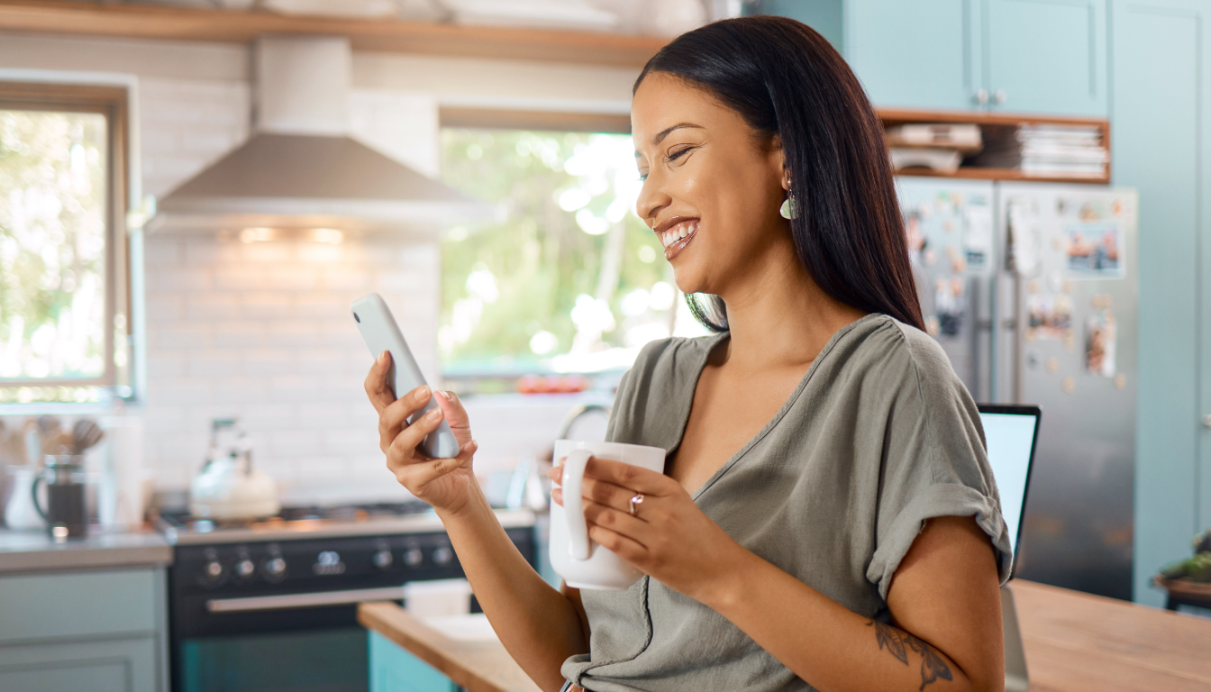 The happy woman is watching something on her smartphone and smiling. She's holding a cup in her hands, with the kitchen blurred in the background.